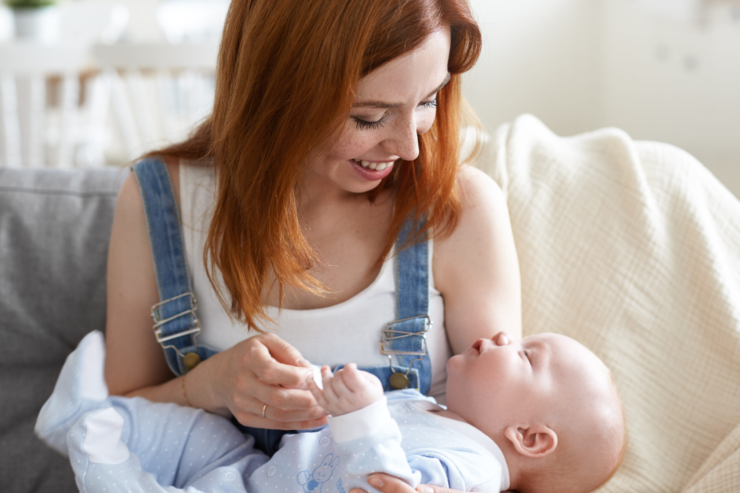 Picture of beautiful charming young red haired Caucasian female wearing denim jumpsuit sitting on couch and holding cute baby, talking or singing to him, touching hands and smiling broadly